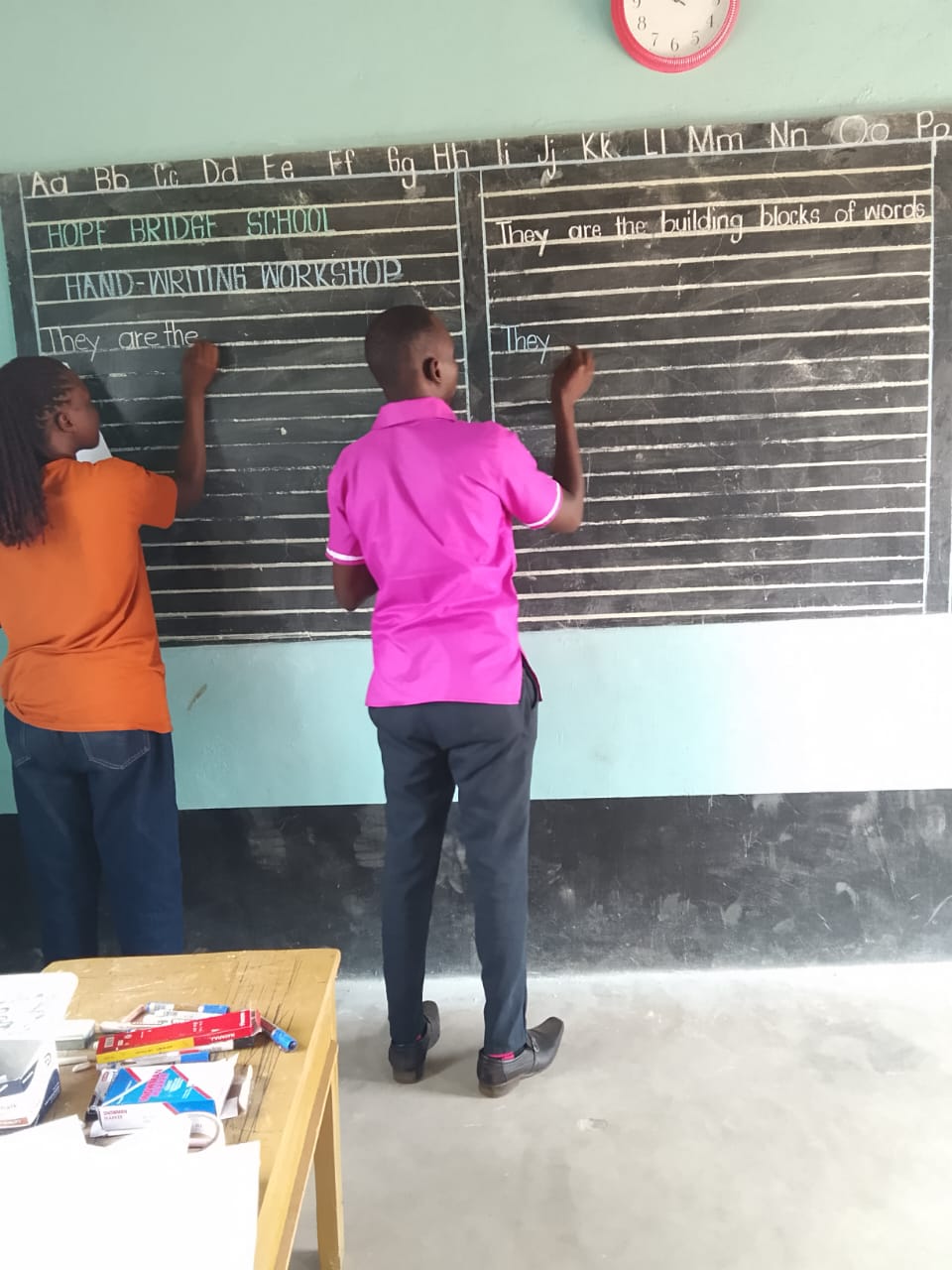 Two students at the chalkboard during Hope Bridge's handwriting workshop, writing They are the building blocks of words